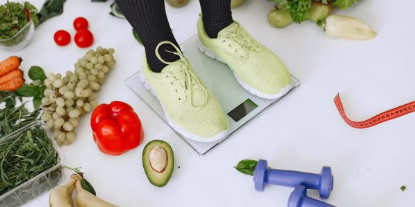 Person standing on scales surrounded by fresh produce and fitness equipment symbolizing a balanced lifestyle.